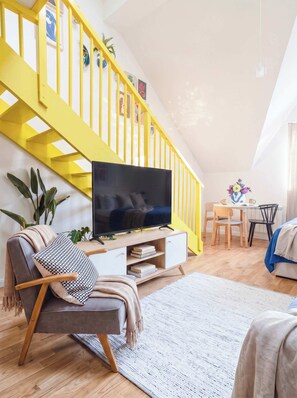 Een woonkamer met een gele trap, een flatscreen televisie op een houten console, een bank, een eettafel met stoelen, en een plant.