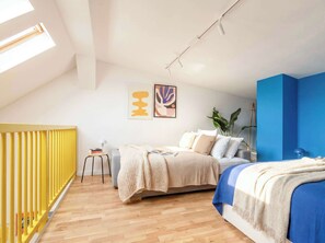 A modern bedroom with a sloped ceiling, a bed with a blue accent wall, a small table with books, and a yellow radiator.