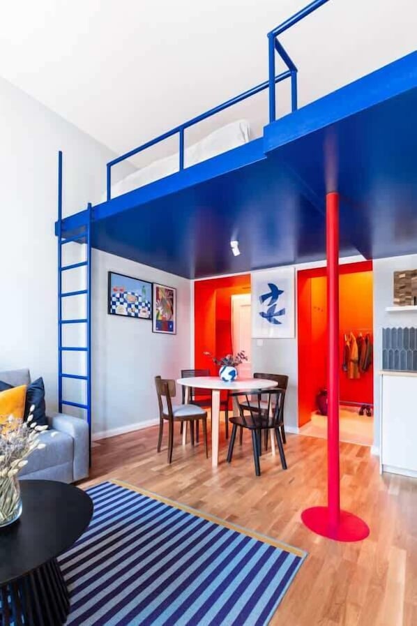 A room with a loft bed, a dining area with chairs, a table, and a red pole.