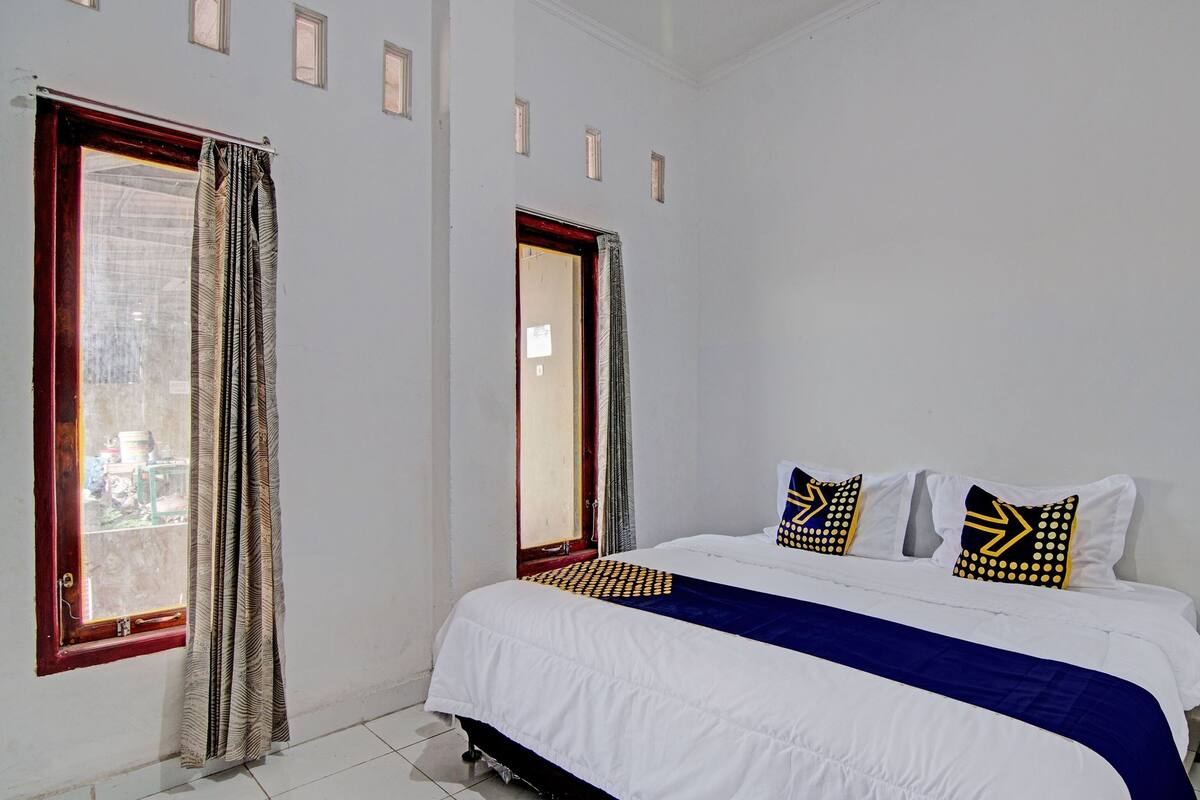 A bedroom with a large bed, two windows with curtains, and a view of the outside.