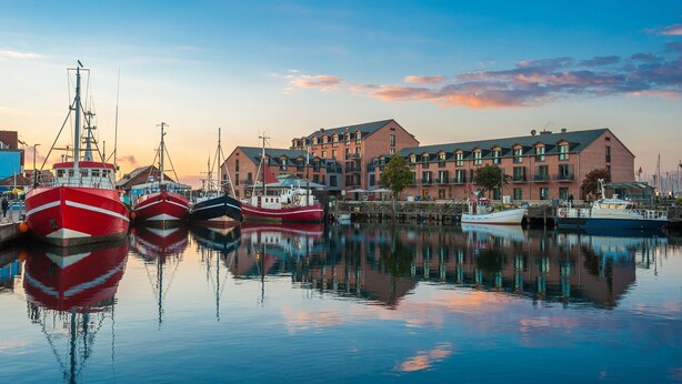 Un port de plaisance où sont amarrés plusieurs bateaux, un grand bâtiment doté de nombreuses fenêtres et de balcons, et une surface d’eau calme qui reflète les bateaux et le bâtiment.