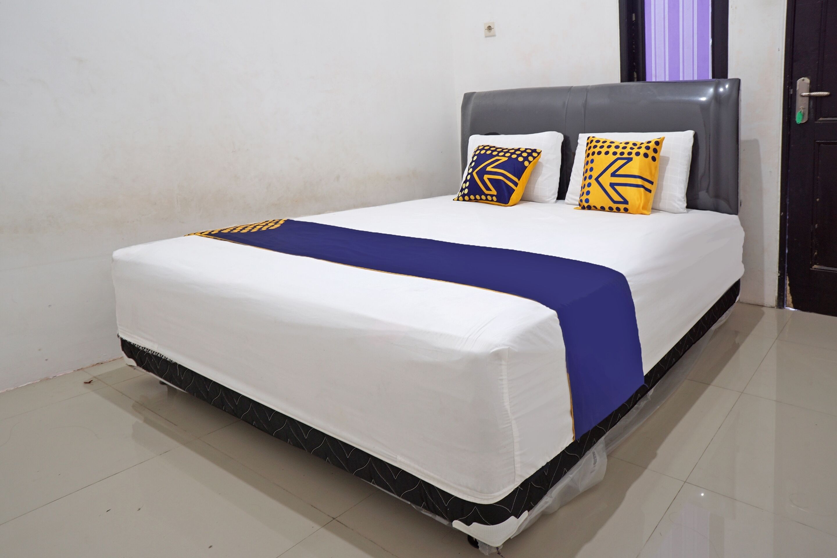 A neatly made bed with a white mattress, blue bedspread, and two decorative pillows with geometric patterns.