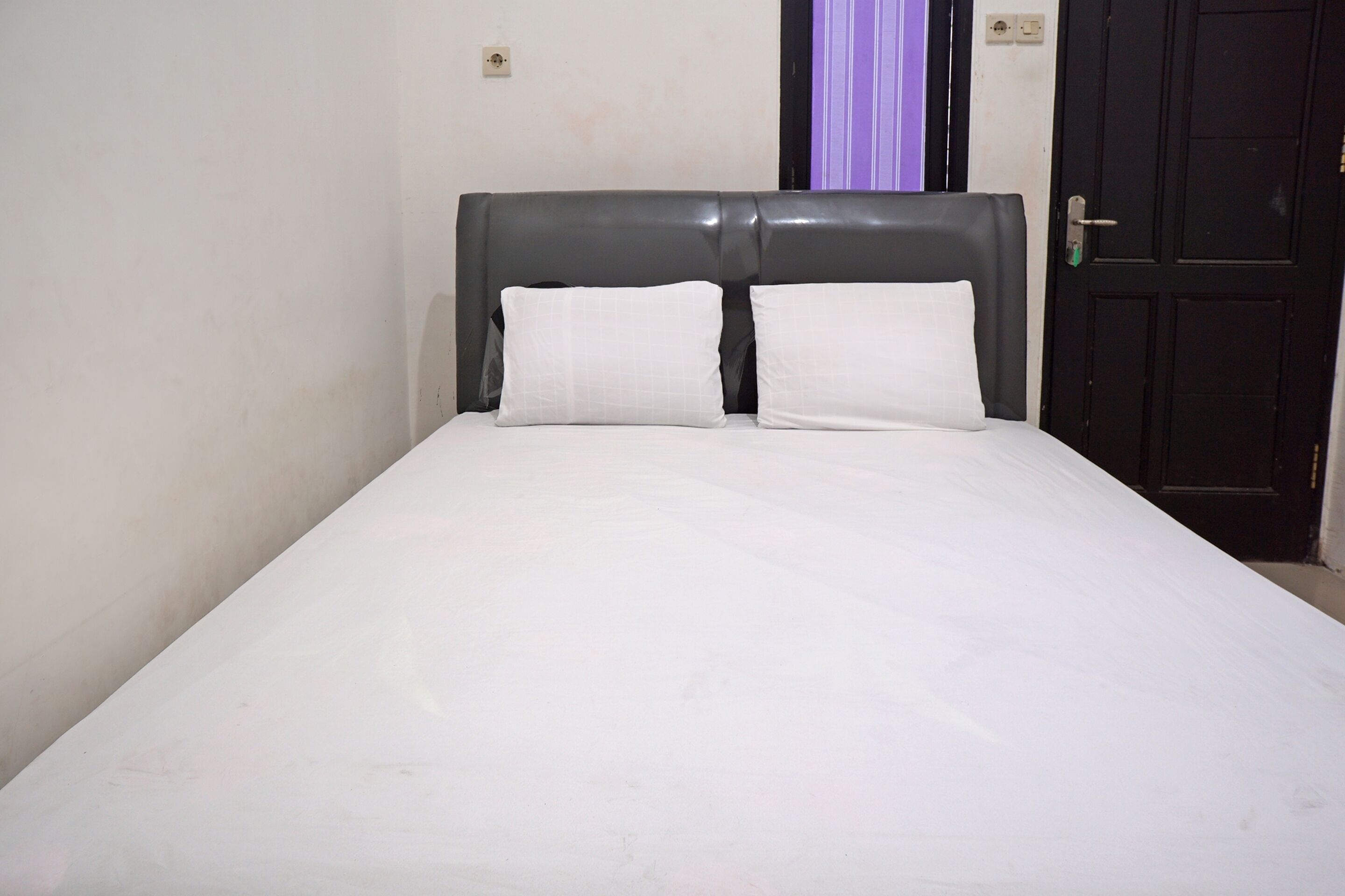 A single bed with a grey headboard and white bedding in a room with plain walls and a closed door.