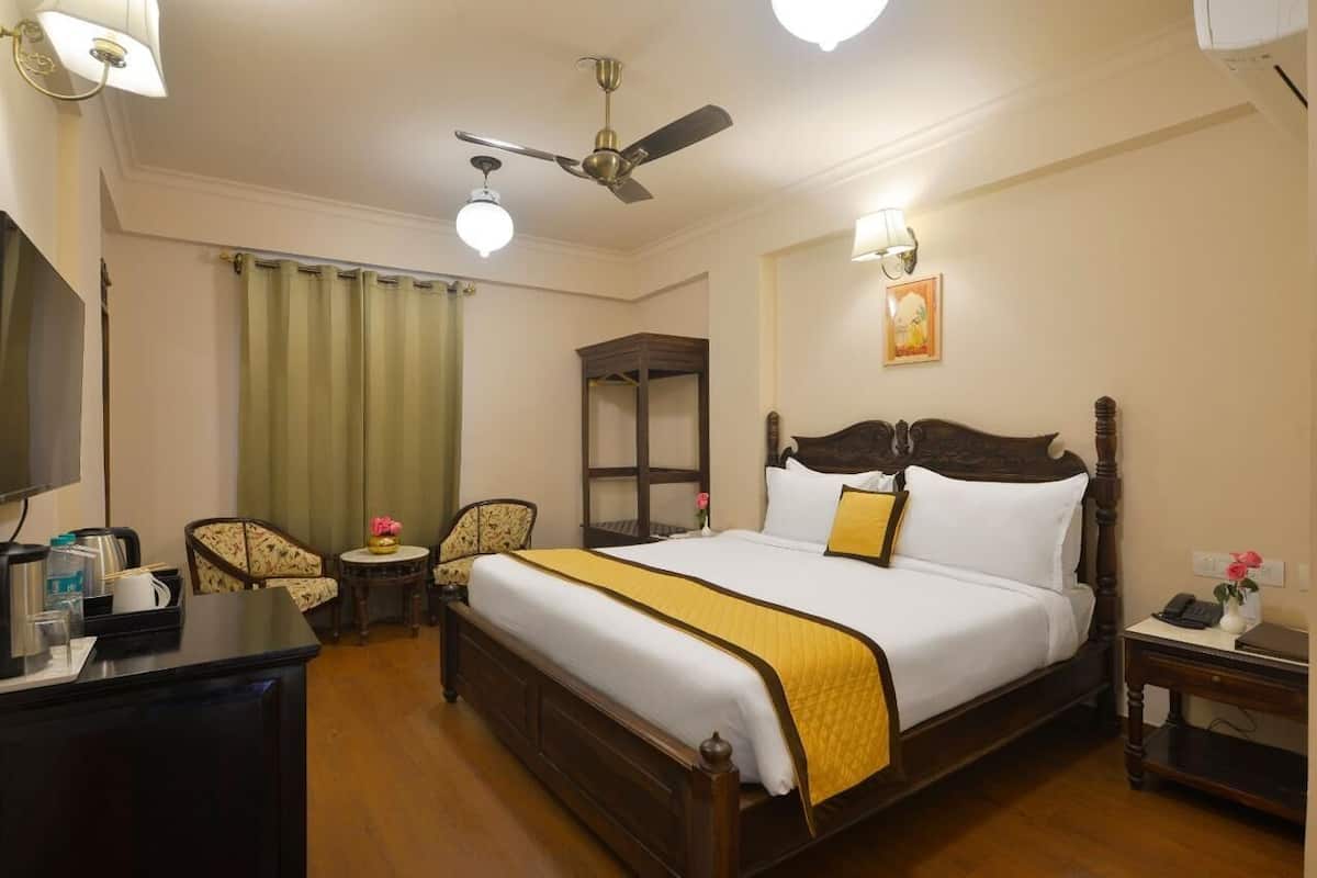 Quarto de hotel com uma cama grande, uma escrivaninha com cadeira, uma mesinha com cadeira, um ventilador de teto e um abajur fixado na parede.