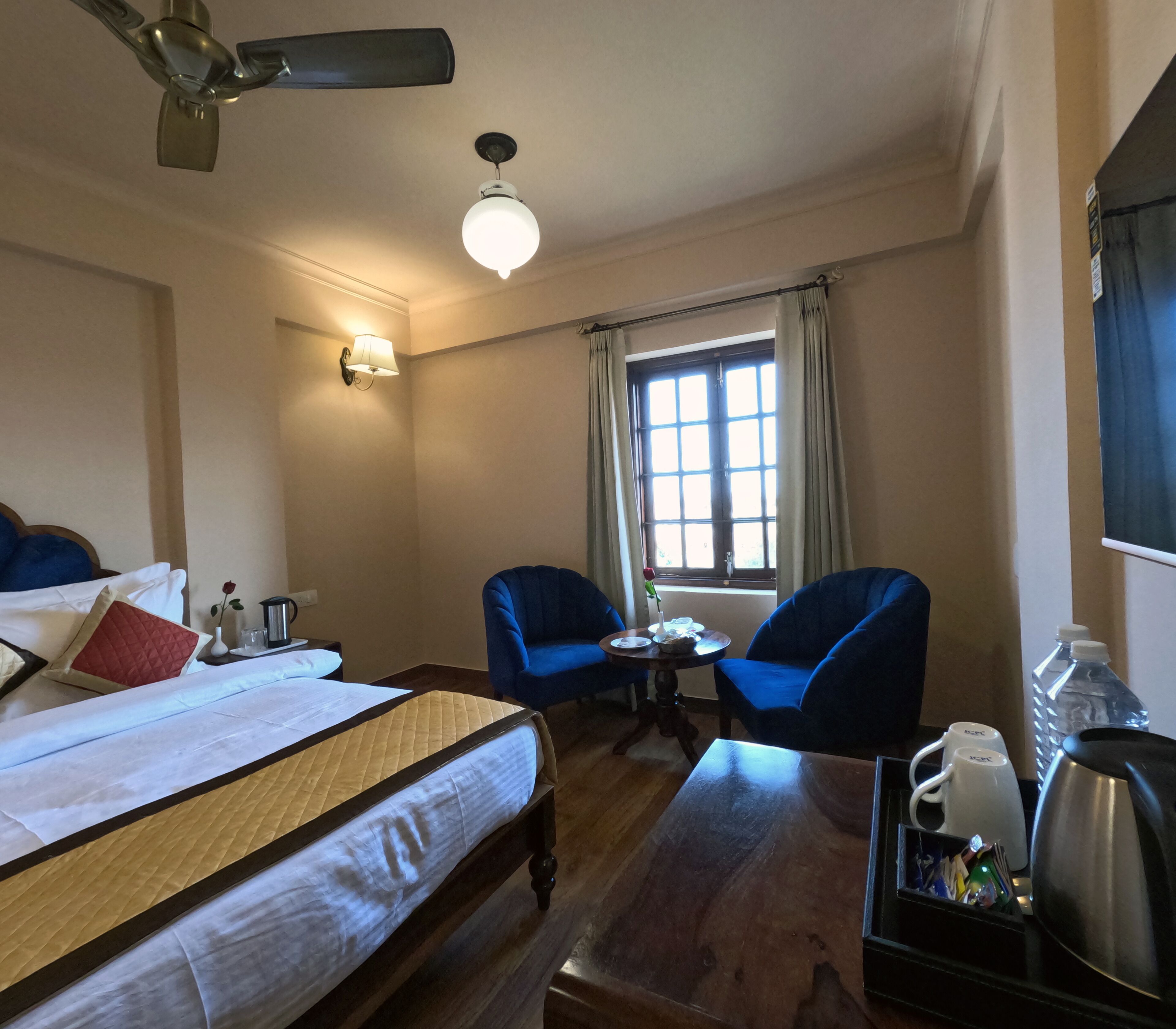 Habitación de hotel con una cama, dos sillones azules, una mesita, una ventana con cortinas, un ventilador de techo y una lámpara de pared.