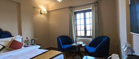 A hotel room with a bed, two blue armchairs, a small table, a window with curtains, a ceiling fan, and a wall-mounted lamp.
