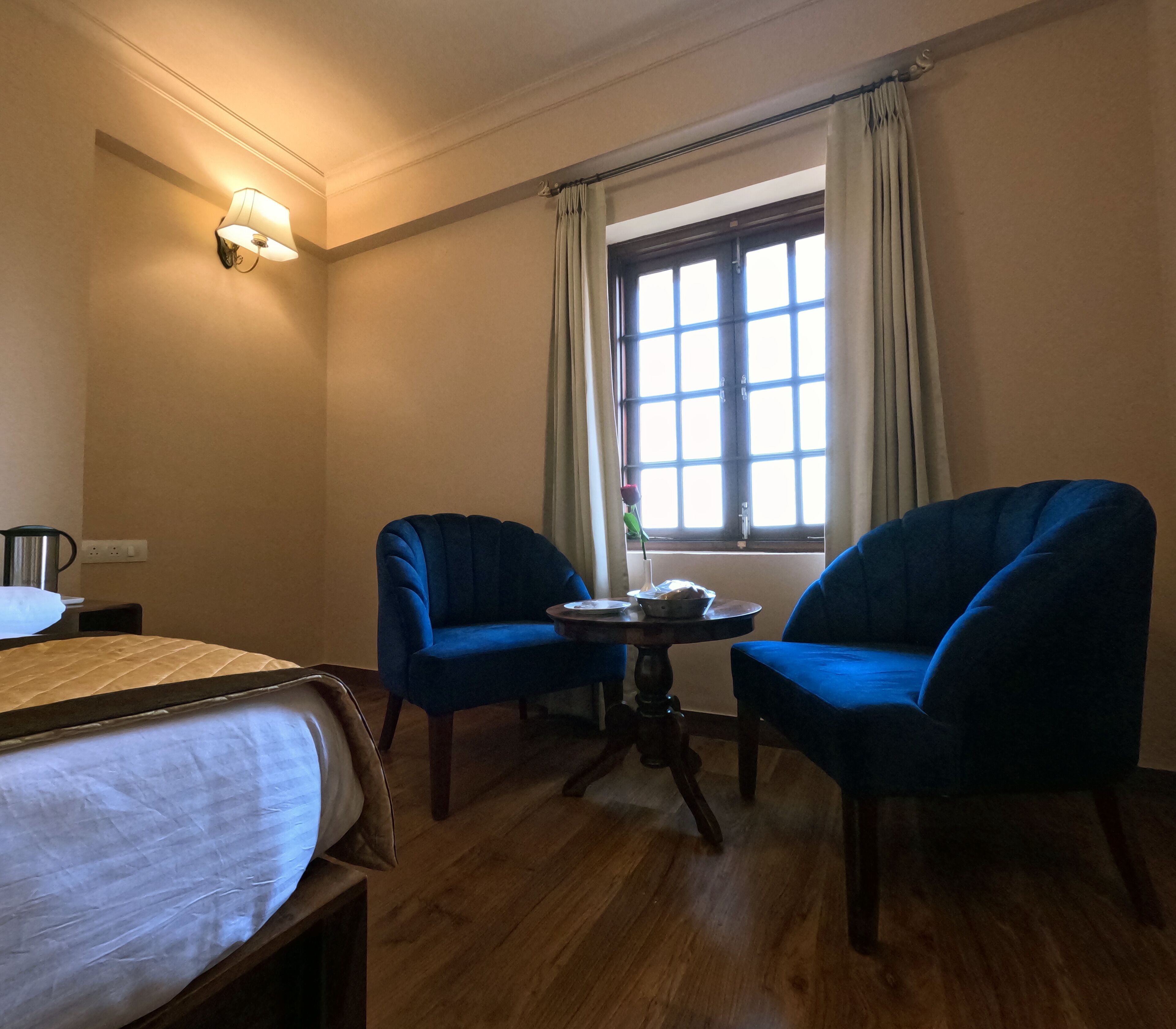 A hotel room with a bed, two blue armchairs, a small table, a window with curtains, and a wall-mounted light fixture.