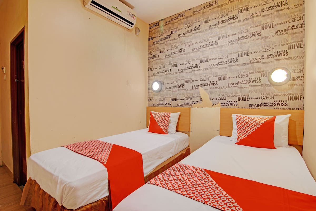 A hotel room with two single beds, each with a red and white patterned bedspread and matching pillows. The room has a beige wall with a word cloud wallpaper and two wall-mounted reading lights.