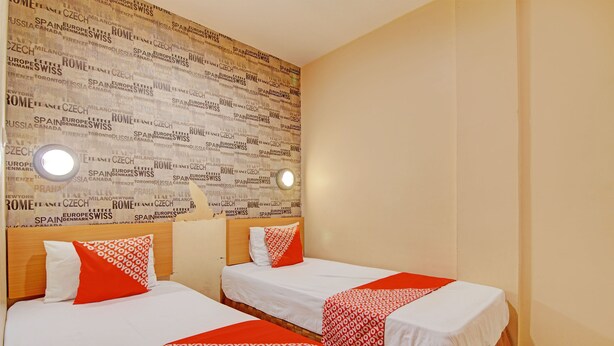 A hotel room with two beds, each with white bedding and red accents, a wooden headboard, and a wall with text in multiple languages.