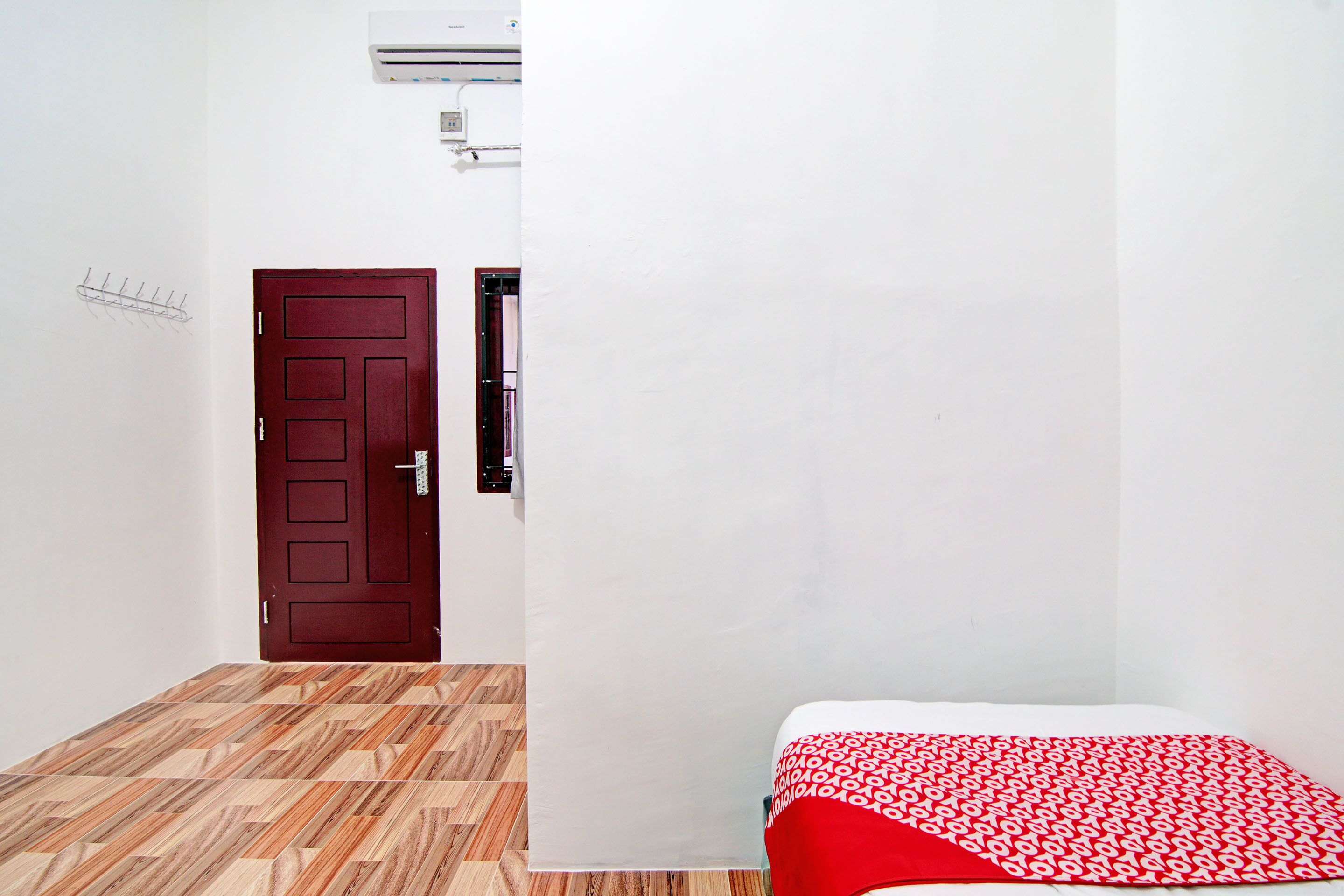Una habitación con suelo de madera, una puerta roja, una cama con colcha de rayas rojas y blancas, y un aire acondicionado instalado en la pared.