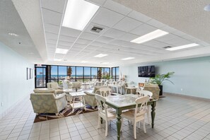 A spacious indoor area with multiple tables and chairs, a large window with a view, and a flat-screen TV mounted on the wall. - Beachfront Getaway at Phoenix 8, 1202 (Orange Beach)