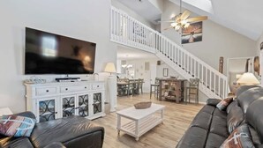 Ein geräumiges Wohnzimmer mit einem großen Flachbildfernseher, einem schwarzen Ledersofa, einem weißen Couchtisch und einer Treppe, die zu einer oberen Ebene führt.
