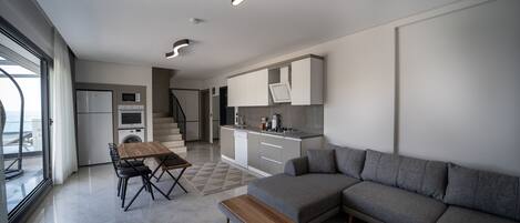 A modern kitchen with integrated appliances, a dining area with a wooden table and chairs, and a grey sofa in a spacious living area with large windows.