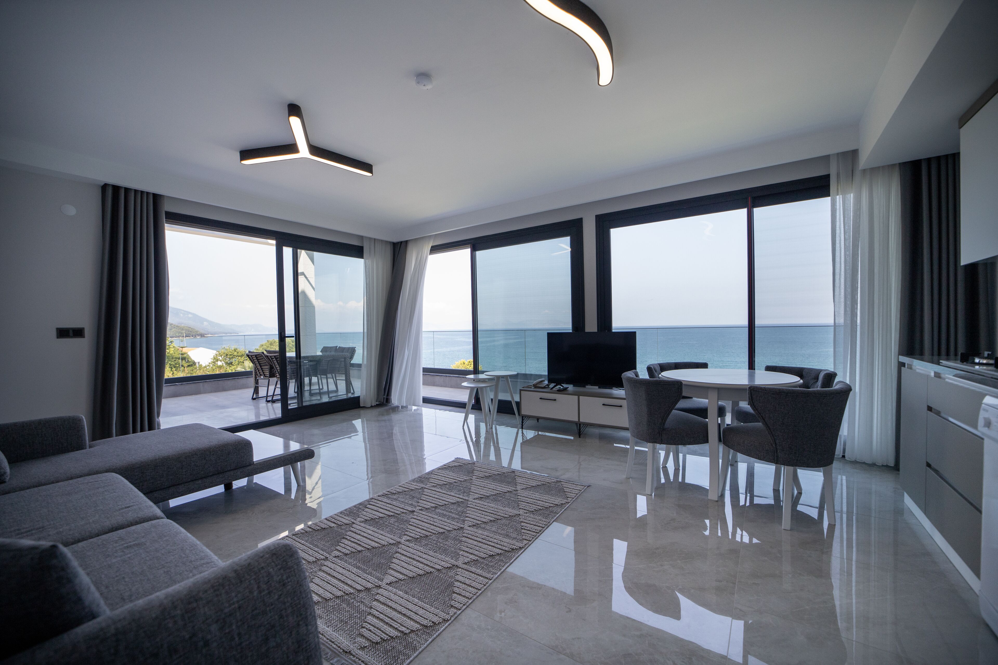 A modern living room with a large window, a flat-screen TV, a dining area, and a kitchen.
