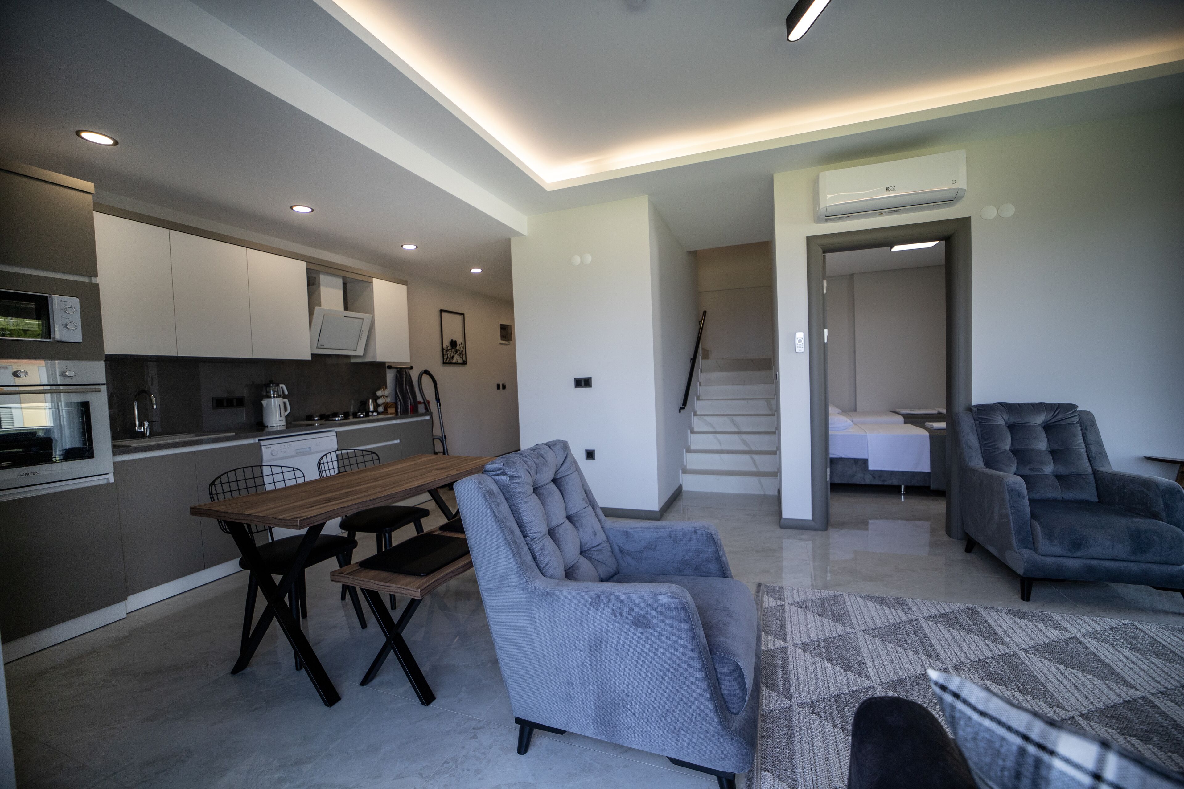 A modern kitchen with built-in appliances, a dining area with a table and chairs, and a living space with a sofa and a staircase leading to a bedroom.