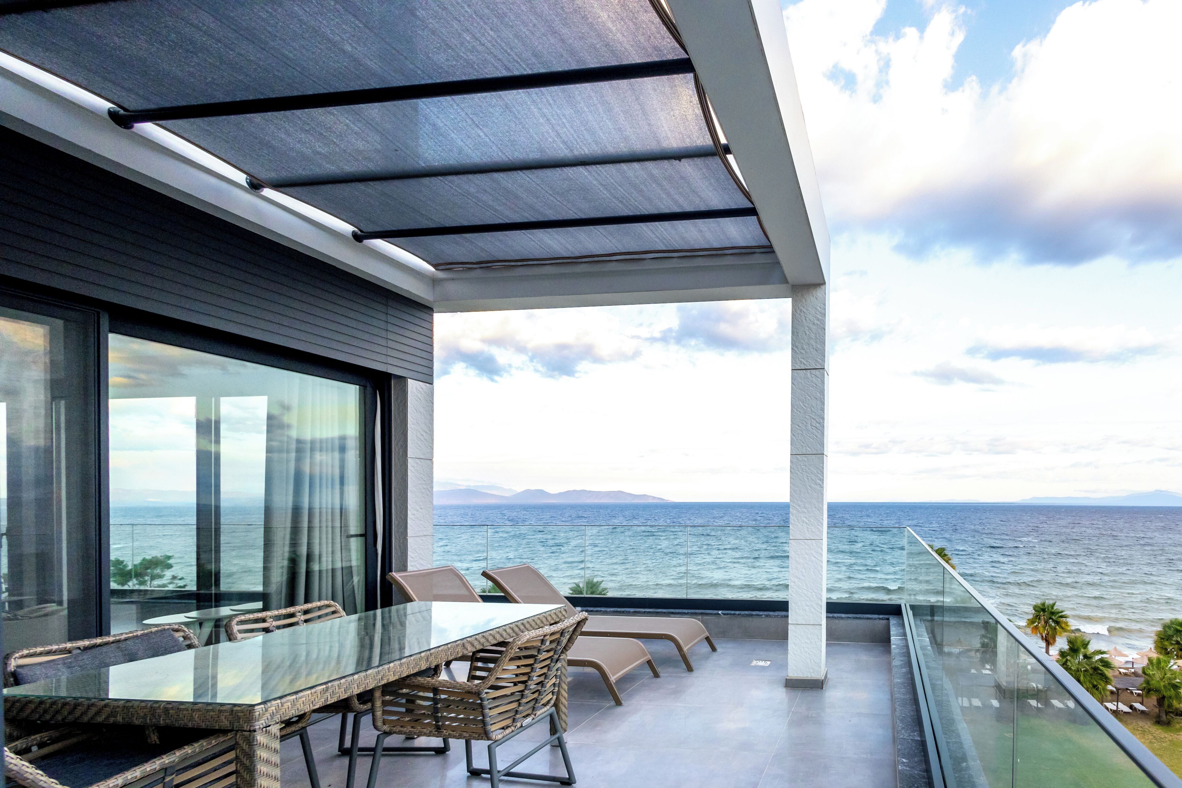 A modern outdoor dining area with a glass table and wicker chairs, overlooking a scenic ocean view.