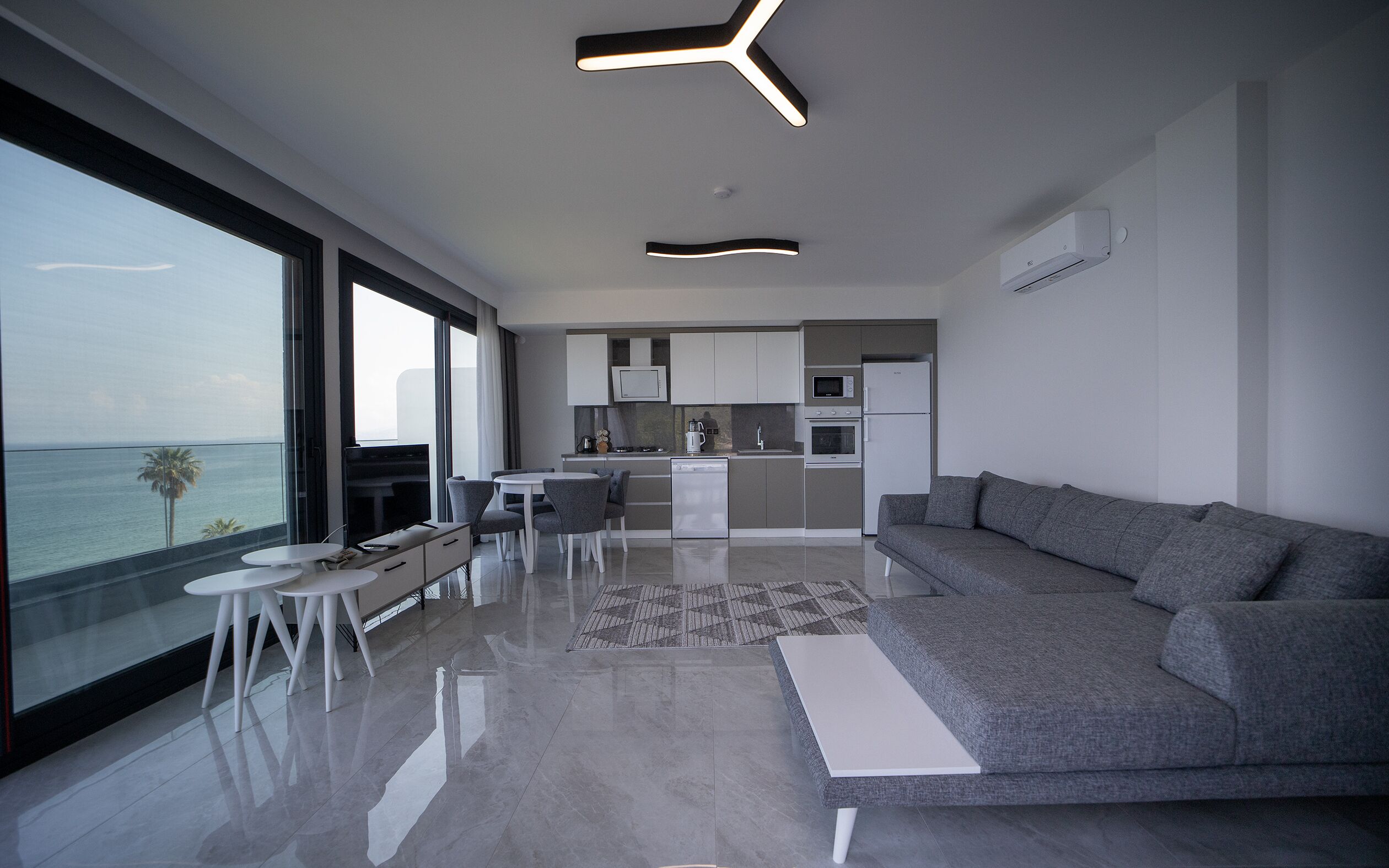 Un salon moderne avec un canapé gris, un coin repas avec une table blanche et une cuisine avec des meubles blancs.