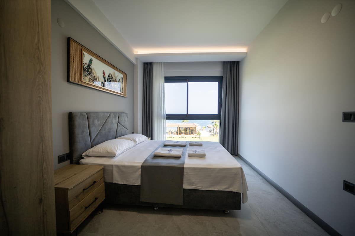 Een moderne slaapkamer met een groot bed, een nachtkastje, een houten dressoir en uitzicht op het strand door een groot raam.