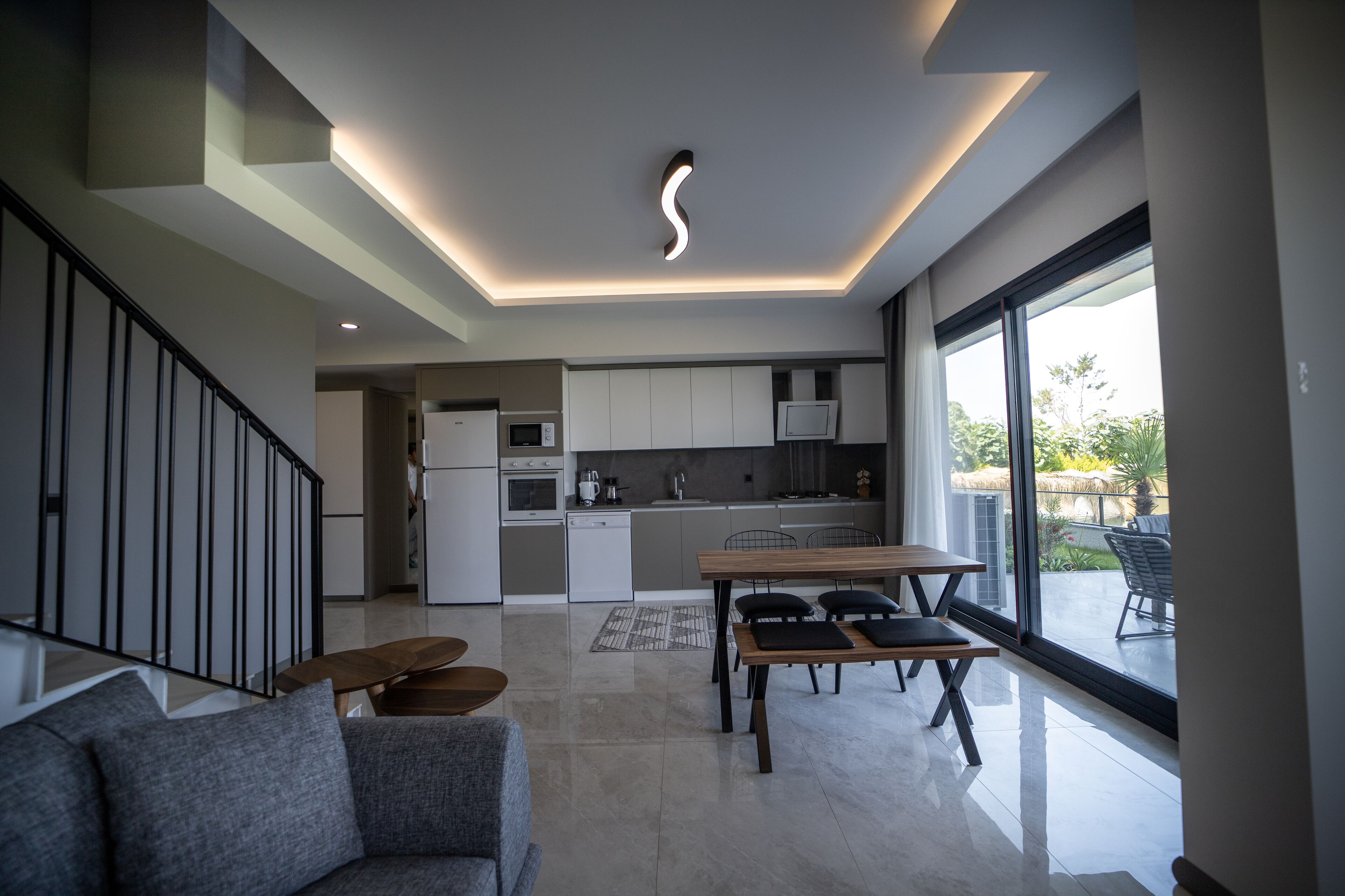A modern kitchen with white cabinets, a dining area with a wooden table and benches, and a large window offering a view of the outdoors.