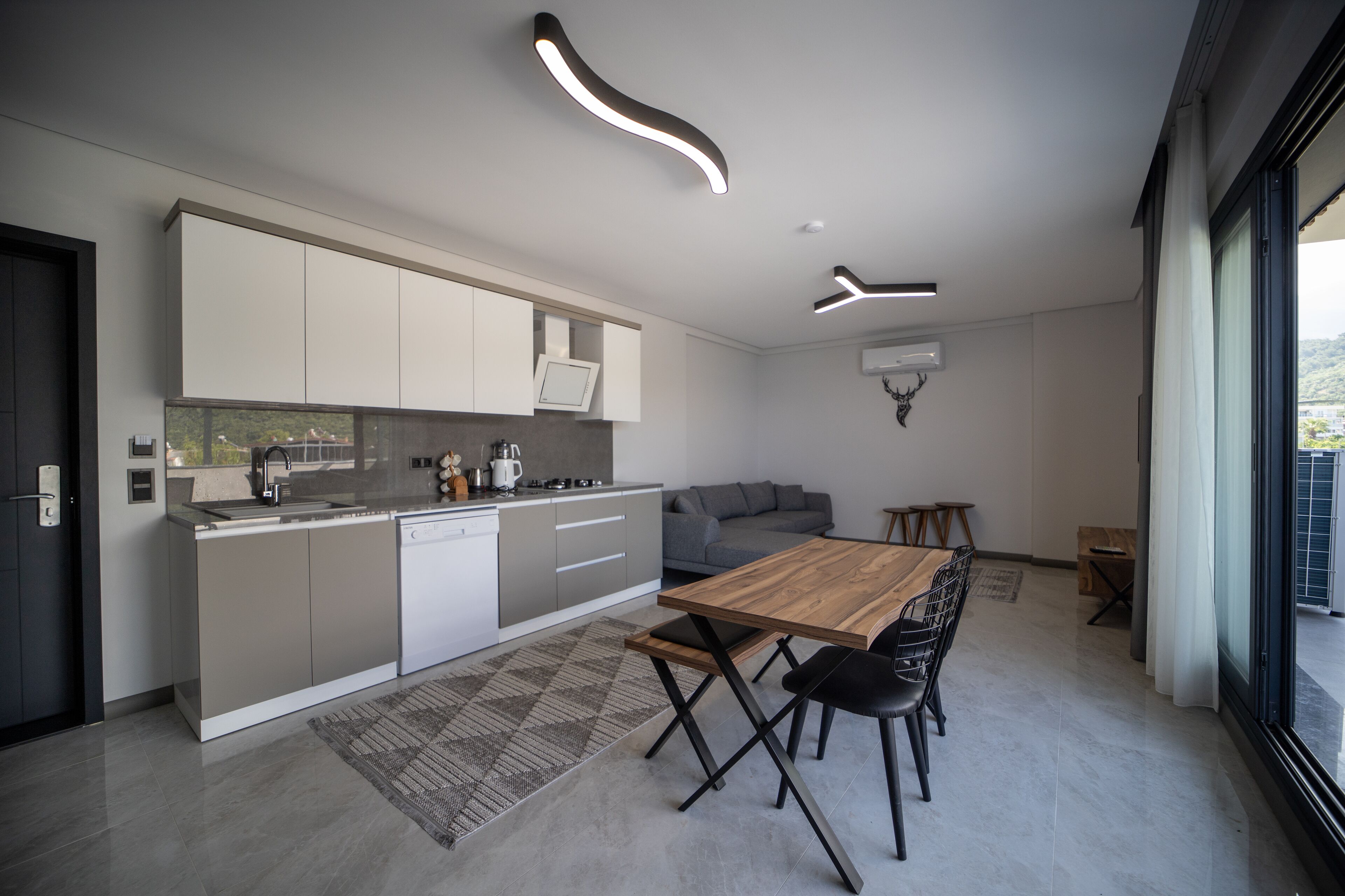 A modern kitchen with a dining area, featuring a wooden table and chairs, a grey sofa, and contemporary lighting fixtures.