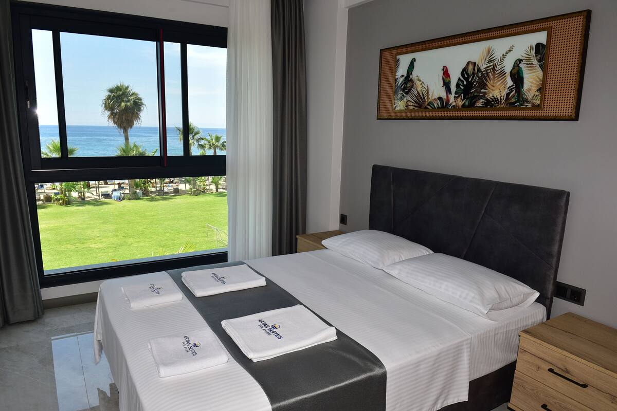 Een hotelkamer met een groot raam, een bed met witte lakens, een hoofdbord, een nachtkastje en een schilderij aan de muur.