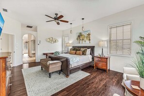 Une chambre à coucher comprenant un lit, une table de chevet, une chaise, un bureau, un ventilateur de plafond, un tableau et une fenêtre avec des stores.