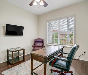 Een kamer met een houten bureau, een leren fauteuil, een televisie aan de muur en een raam met jaloezieën.