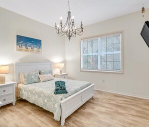 A bedroom with a bed, nightstands, a chandelier, a window with blinds, and a beach-themed painting. - Luxury 6BR Retreat with Beach Access (Sarasota)