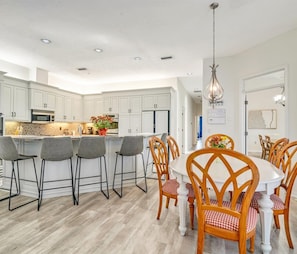 A modern kitchen with white cabinets, a central island, and bar stools. There is a dining area with a round table and wooden chairs. - Luxury 6BR Retreat with Beach Access (Sarasota)