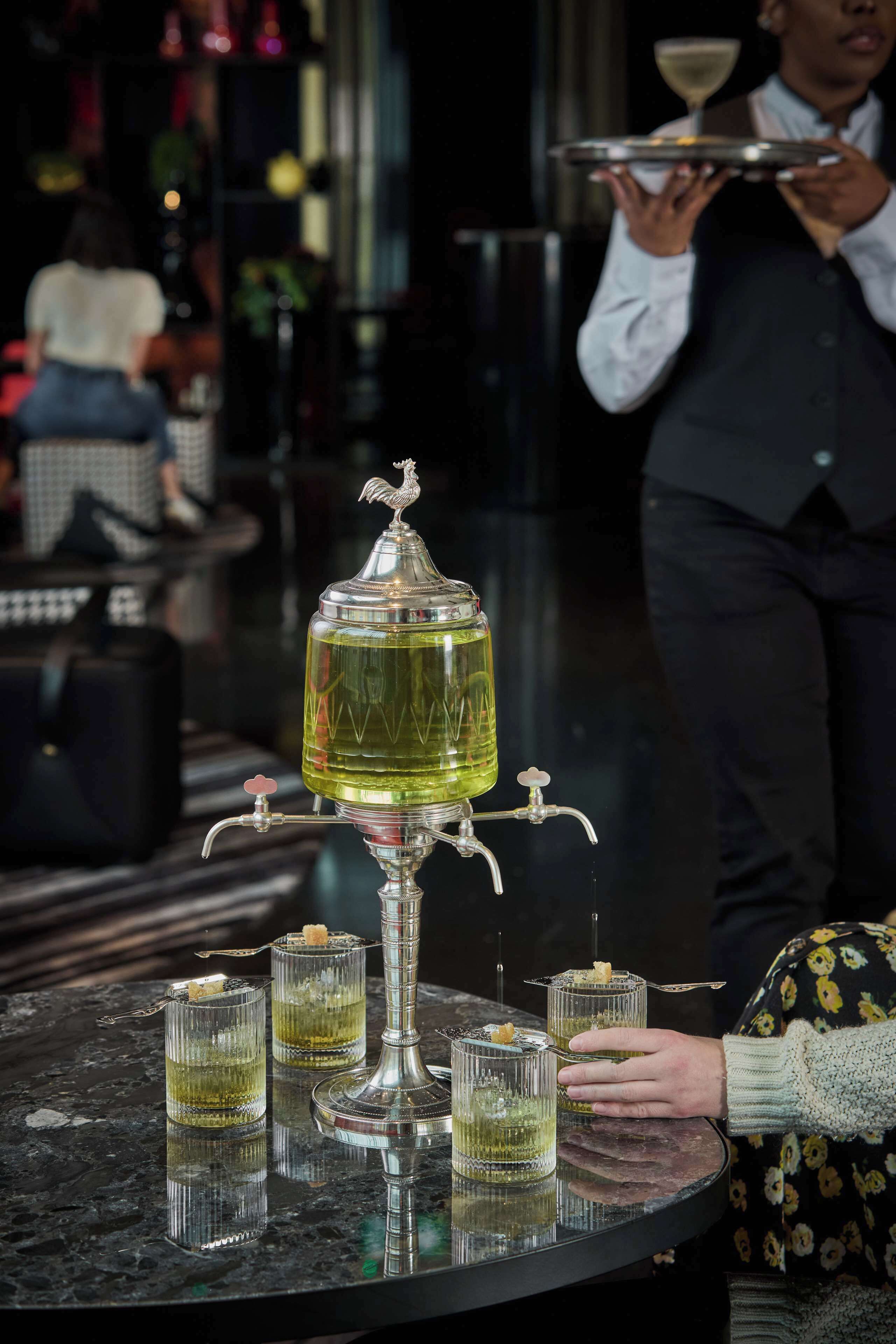 A bar area with a central liquor dispenser and several glasses of a yellowish beverage.