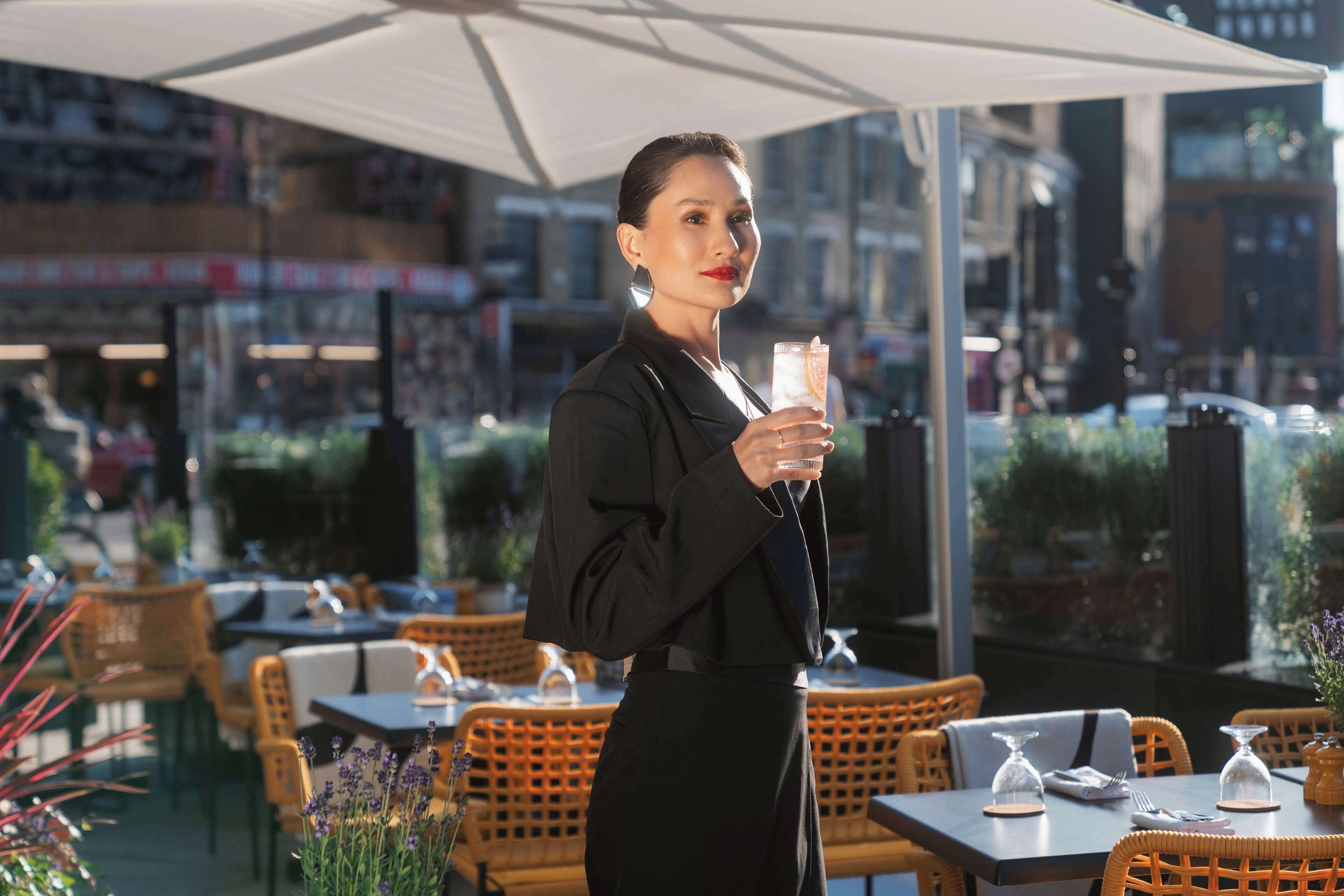 En person står ved en udendørs café med fletmøbler og parasoller, mens vedkommende holder et drikkepål.