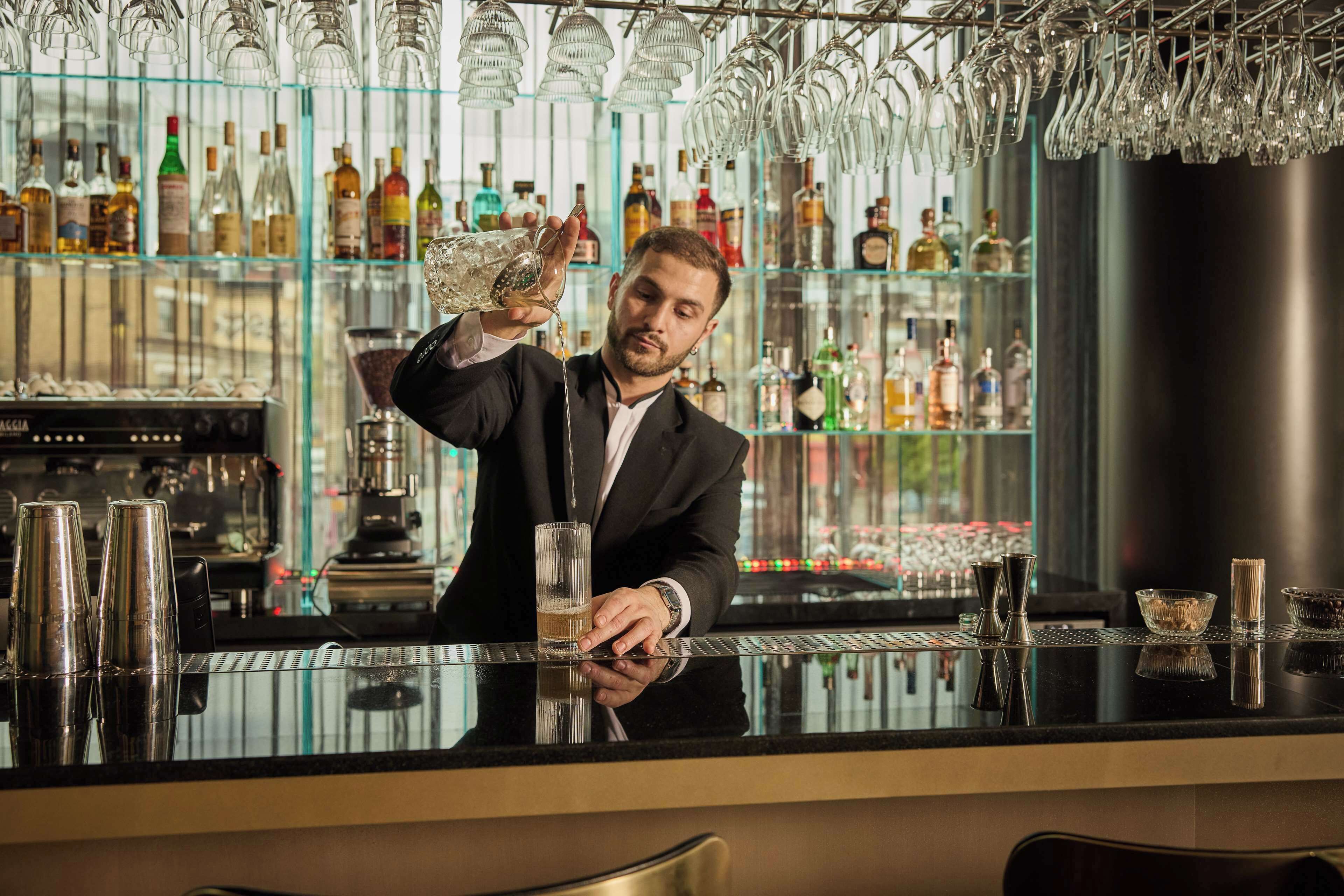 Un bar con un cantinero sirviendo una bebida. La barra tiene un estante de vidrio con diversas botellas y un recipiente negro grande.