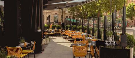 An outdoor cafe with tables and chairs, a glass enclosure, and a view of the street.