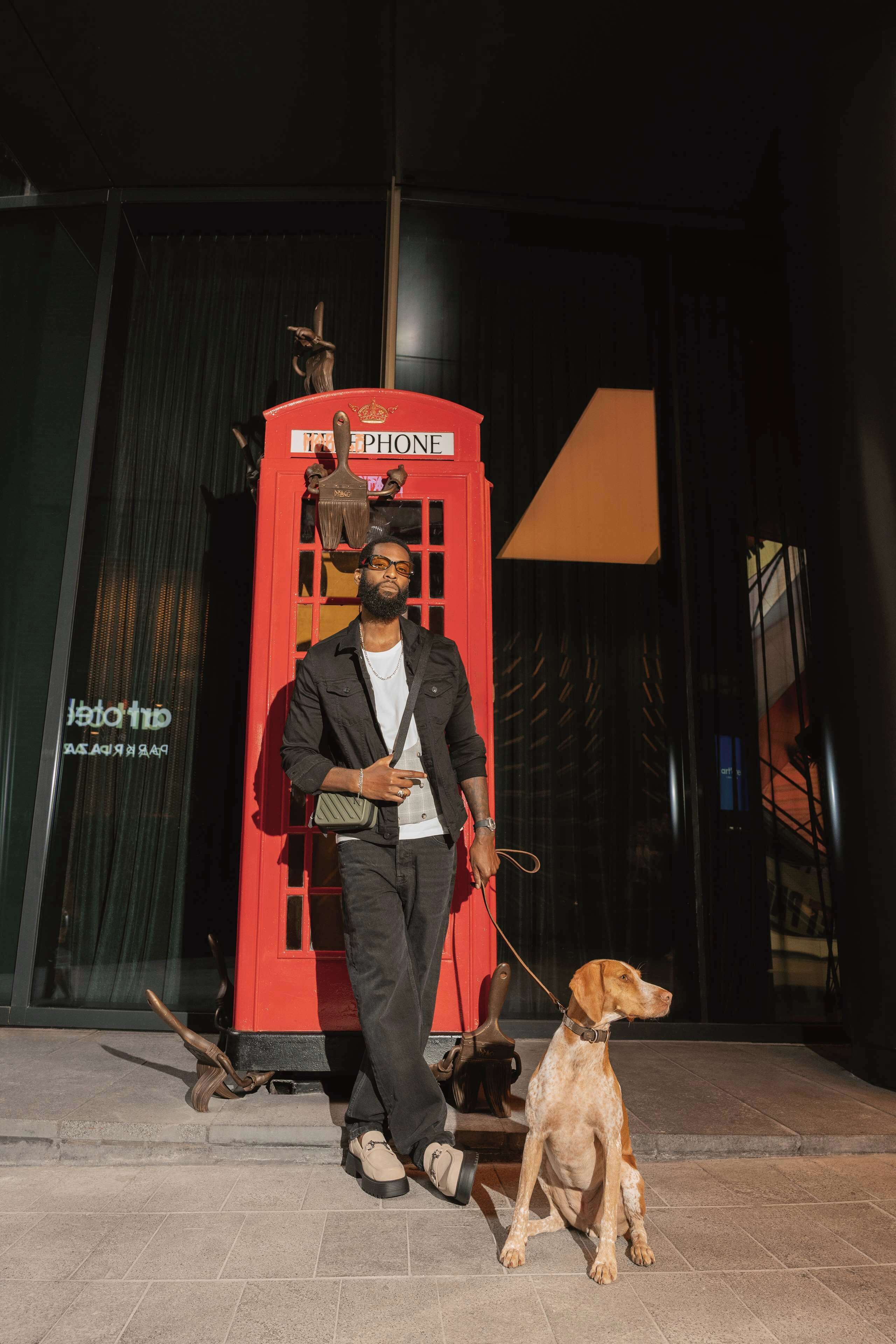 Une personne se tient à côté d’une cabine téléphonique rouge, et un chien est tenu en laisse à ses côtés.
