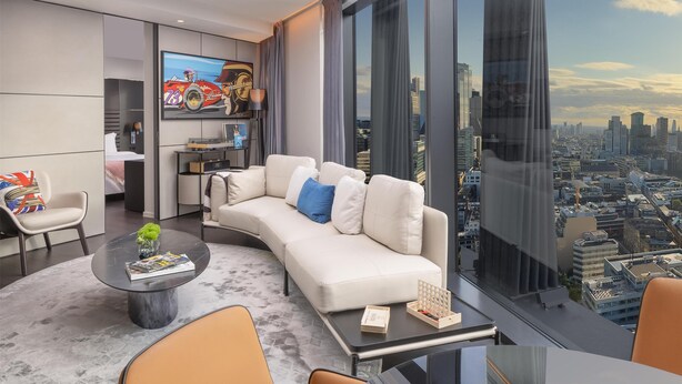 A modern hotel room with a large window offering a city view, a white sofa, a coffee table, and a flat-screen TV mounted on the wall. - art'otel London Hoxton (London)