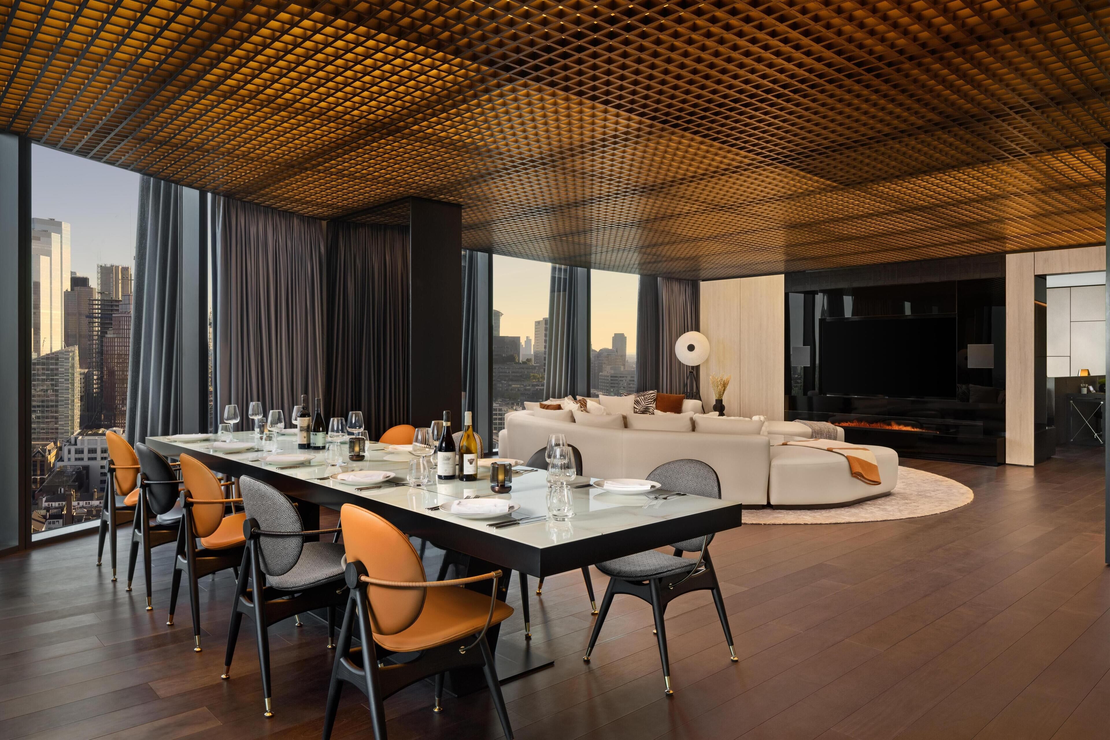 A modern dining area with a long table set for six, surrounded by chairs. A large window offers a city view.