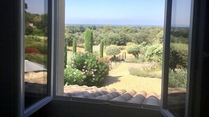 A view through a window with a clear sky, a garden with various plants, and a distant landscape. - Villa Rêves de Sorgues (Saumane-de-Vaucluse)