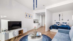 Ein modernes Wohnzimmer mit Flachbildfernseher, Glastisch, Sofa und einem Bett mit blauer Bettwäsche.