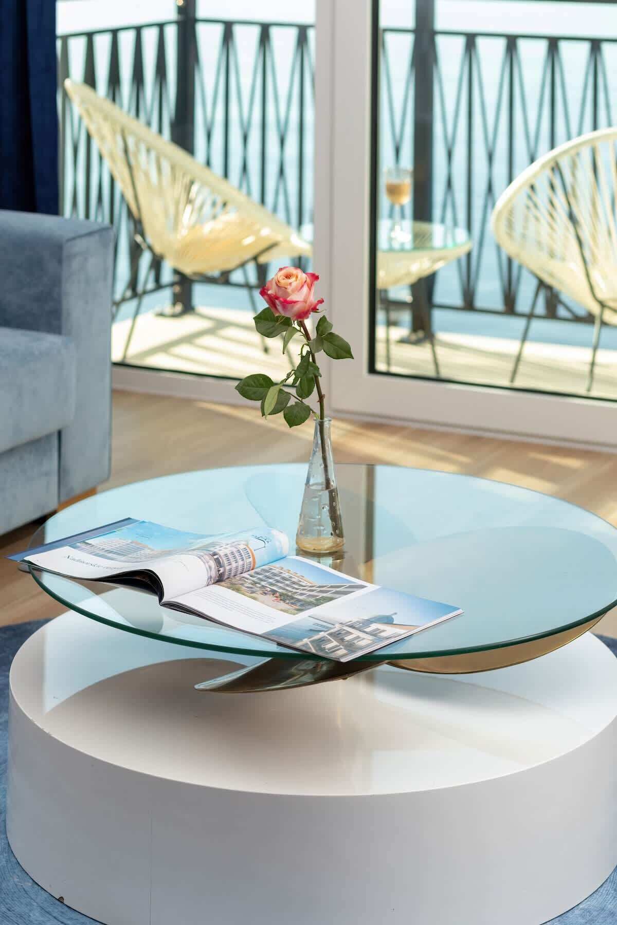 A modern living room with a glass coffee table, a vase with a single rose, and a magazine.