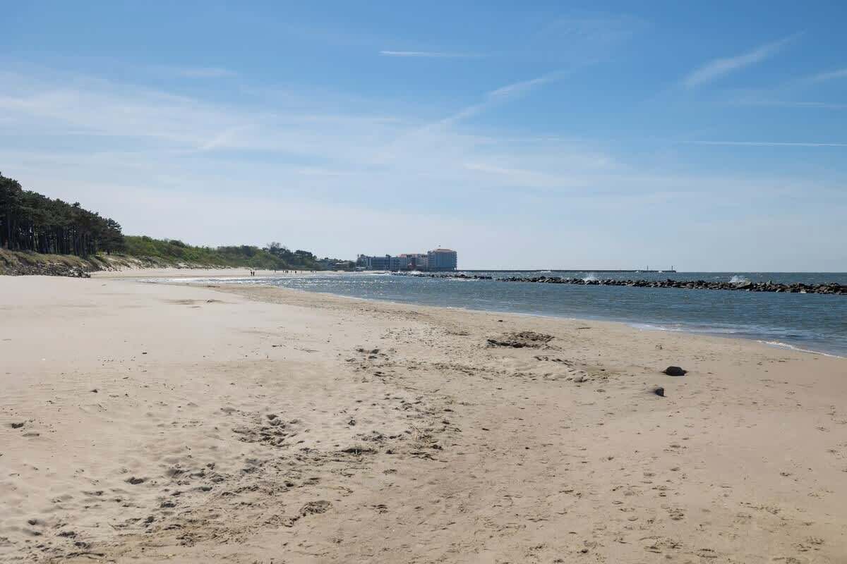 Una amplia playa arenosa con agua tranquila, un rompeolas rocoso y un edificio a lo lejos.