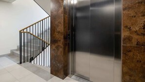 A modern elevator with a metallic finish, adjacent to a staircase with a wooden handrail and black railings.