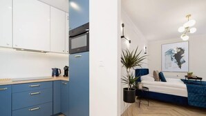 Een moderne kamer met een keukenhoek, met blauwe kastjes, een magnetron en een koffiezetapparaat. Er staat een bed met een blauw hoofdeinde, een nachtkastje en een potplant.