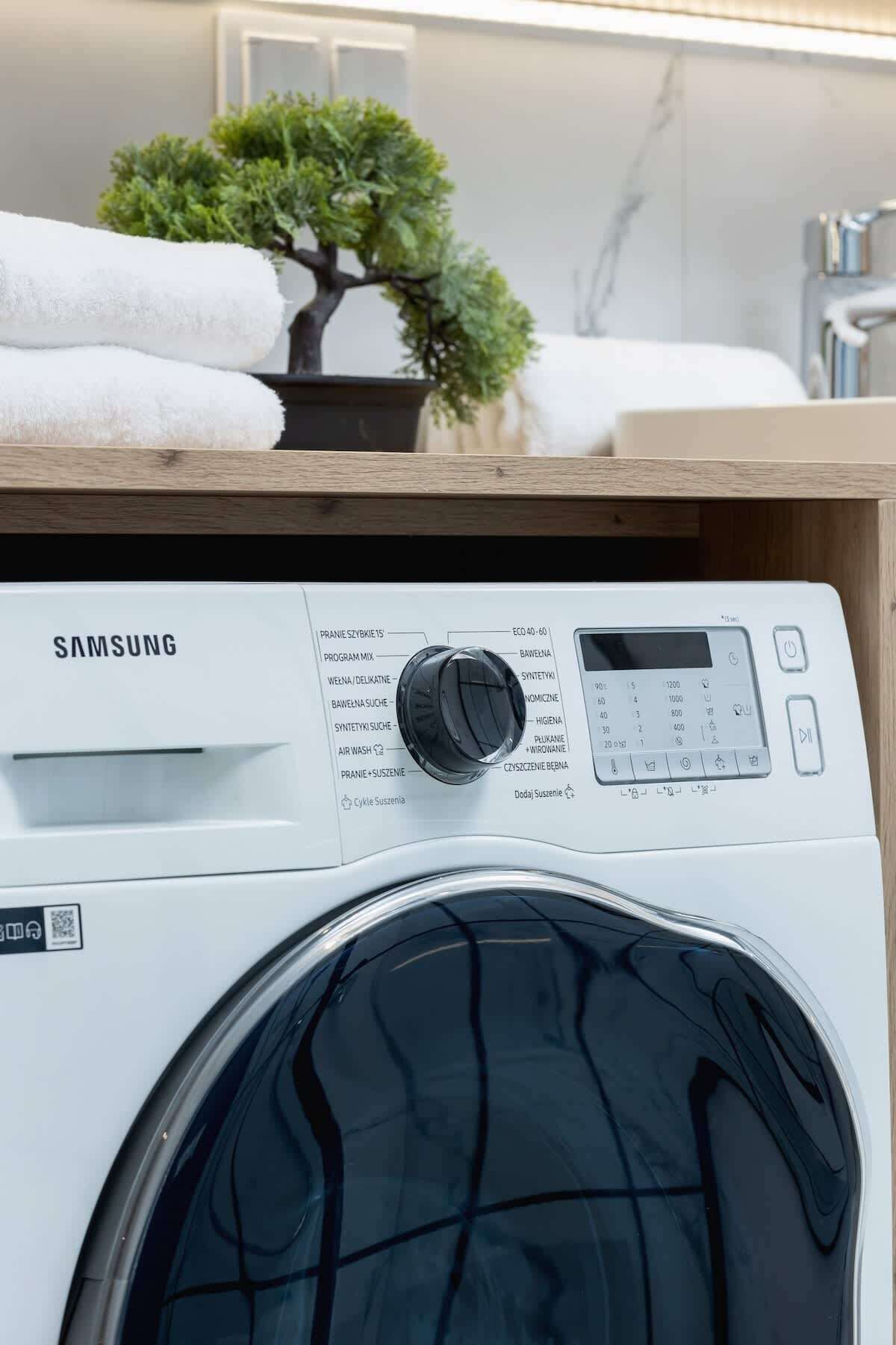 Una lavatrice Samsung con un pannello di controllo, una piccola pianta in vaso e una pila di asciugamani bianchi sopra.