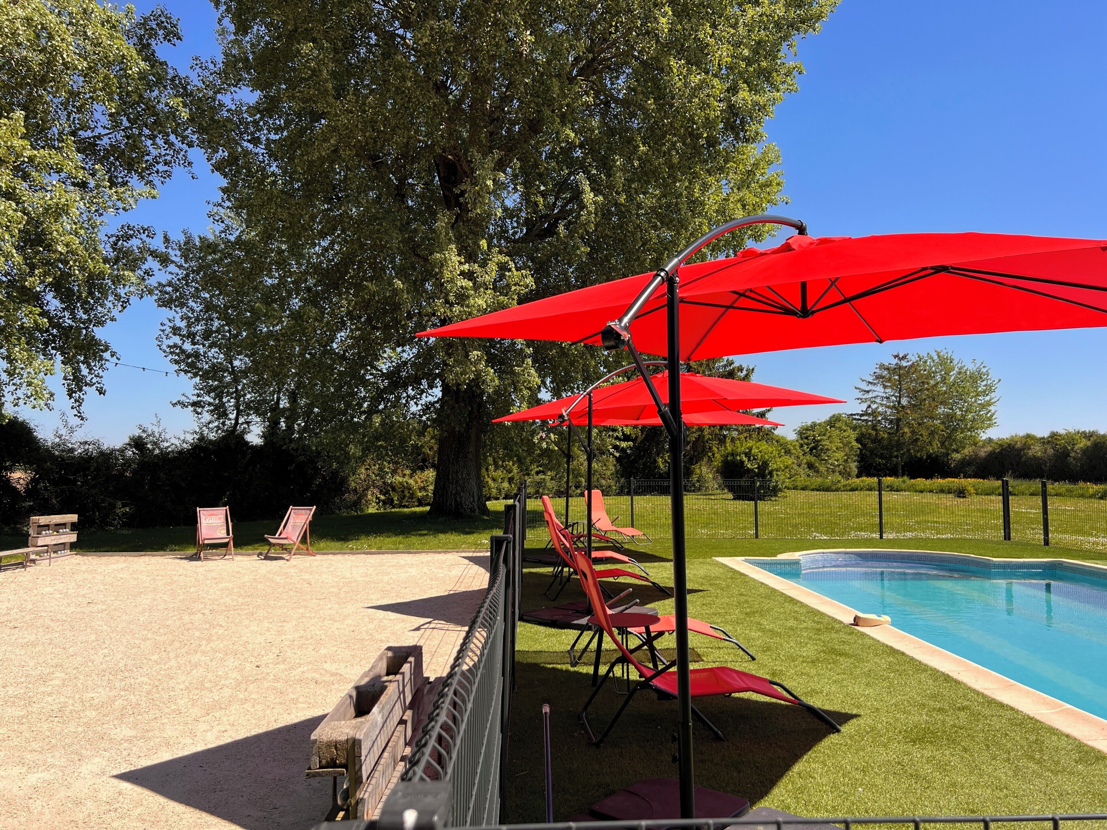 A pool area with red umbrellas, a pool, and a grassy area with chairs.