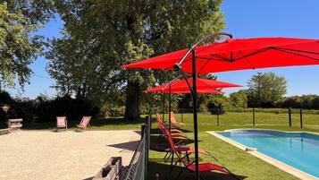 A pool area with red umbrellas, a pool, and a grassy area with chairs.