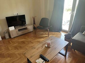 A living room with a flat-screen TV on a wooden stand, a grey armchair, a coffee table with a candle, and a sofa.