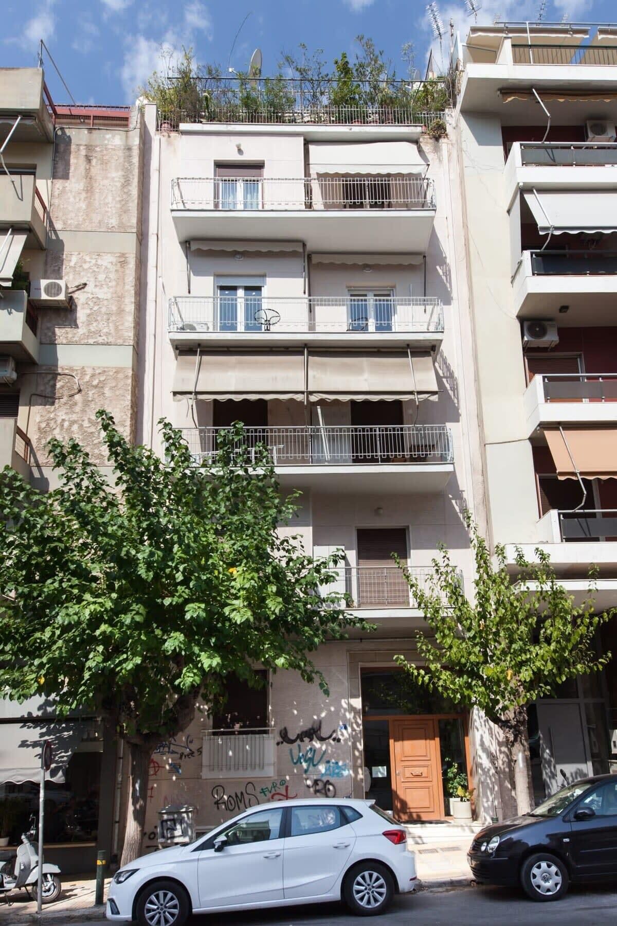 A multi-story building with balconies, a tree in front, and parked cars.