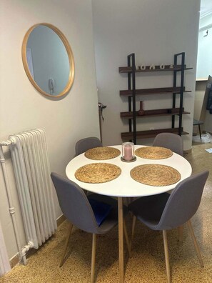 A round table with four chairs, a white radiator, a round mirror, and a wooden shelf unit against the wall.