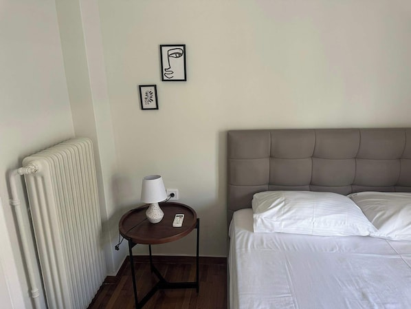 A hotel room with a bed, a round table with a lamp, and two framed artworks on the wall.