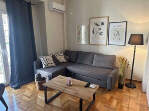 Uma sala de estar moderna com um sofá cinzento, uma mesa de centro de madeira e um abajur. Há obras de arte emolduradas na parede e um cesto no chão.